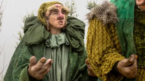 A costumed performer dressed as a troll-like character stands on an outdoor stage. The character wears layered dark green robes with oversized padded shoulders, large sculpted hands, and theatrical facial prosthetics. A second costumed performer is partially visible at the right edge, showing only a green textured shoulder and sleeve. The background includes low greenery and a pale, overcast sky.