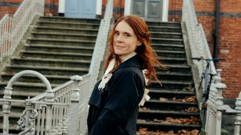 Ellius Grace A woman with long red hair stands in front of a broad outdoor staircase with ornate white railings, set against a red-brick building. She is turned slightly toward the camera and wears a dark coat with light-coloured ribbon details. Fallen leaves are scattered on the steps.