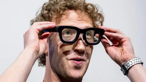 Meta boss Mark Zuckerberg - puts on thick rimmed meta smart glasses. Zuckerberg has medium length blond curly hair and has visible stubble.