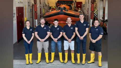 RNLI Portishead Six people - one woman and five men - stand at the entrance to a lifeboat station with an orange lifeboat in the garage behind them. They are smiling to camera and all wearing blue polo shirts with the RNLI logo on them and yellow wellington boots
