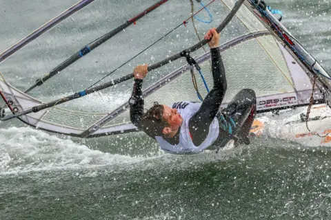 Bill McCandlish A brown-haired man in a wet suit holding onto a sail boat while falling half into the water