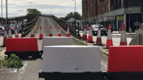 Red and white cones and bollards are blocking off the entrance to the flyover. Cars and traffic lights are in the picture.