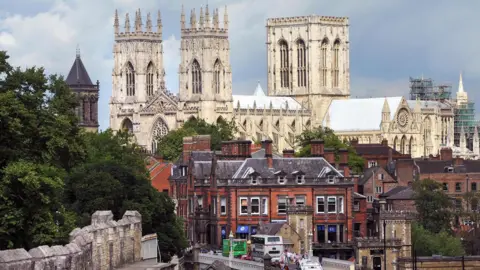 Shot of York Minster, taken from afar. The building is made of stone, which looks white. The rest of York can be seen in front of it. 