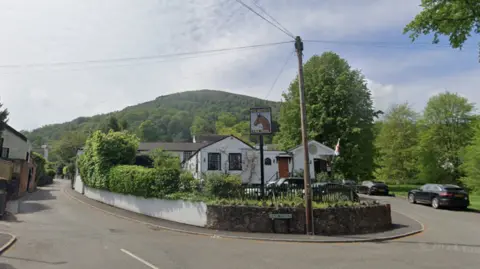 A white pub with a sweeping bend that surrounds it. There is a large hedge to the left of the pub with a sign in the centre of its garden depicting a horse's head. In the background is a large hill covered in woodland.