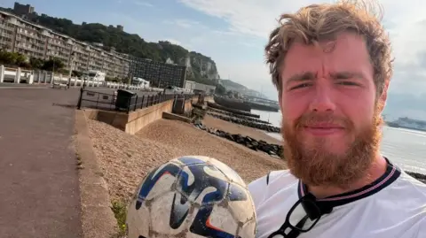 James Lewis is pictured holding a football. He is standing by the coast with a beach and buildings opposite. He has a moustache and beard and wearing a white England top. 