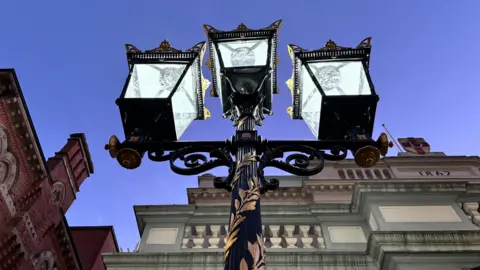 The photo is taken from below the street lamp. It is black with gold leaves engraved on it. The lamp has three lights at the top of it.