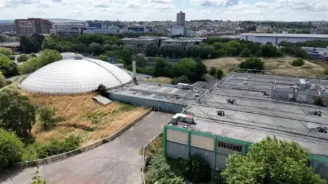 An aerial view of a building with a large white dome surrounded by overgrown grass and hedges.