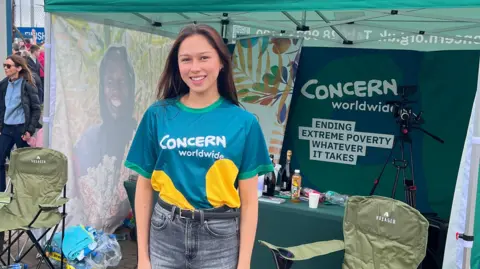 Angela Aumonier lleva una camiseta azul de "Concern Worldwide" y está de pie frente a una carpa benéfica. Tiene el pelo largo y castaño y sonríe.