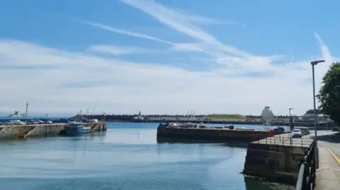 A long industrial pier in Douglas Harbour stretching out in the distance with a boat moored up next to it on a a sunny day.