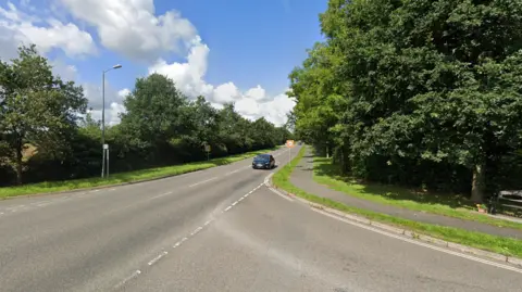 Single laned road with a black car approaching. There are large trees running either side of the road with a footpath on the right-hand side.