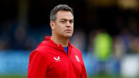 Bath director of rugby Johann van Graan looks on before a Prem Rugby fixture. Van Graan is wearing a red top, which features the logo of the Bath rugby team.