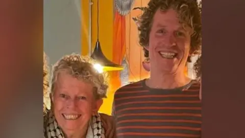 Dale Ryan A man and woman smiling linking arms standing next to each other. The man is on the right and is wearing a dark grey and orange jumper, and has curly dark hair. The woman has short grey curly hair.