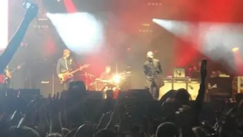 BBC Oasis performs on a stage as fans hold their hands up in the air.