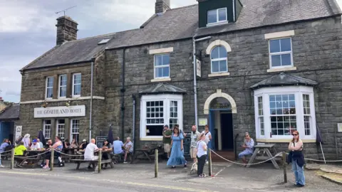 BBC/ Carla Fowler A stone‑built hotel with a sign reading “The Goathland Hotel” stands behind an outdoor seating area where people sit at wooden picnic tables and gather near an arched entrance marked “Hotel Entrance & Family Rooms”. 