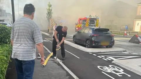 Smoke fills a street, with two fire vehicles in the background parked next to a row of homes. A car is parked on the side of the street next to a freshly installed cycleway with bike signs painted on it. A firefighter in black trousers and t-shirt uses a metal pole to access a fire hydrant. A man with dark hair in a grey striped t-shirt and blue jeans looks on.