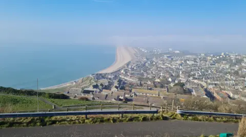 The Klonk A photo taken from an elevated position on top of Portland looks out over the town and towards Weymouth that is shrouded in fog. The rest of the county has vanished into the clouds. In the foreground you can see homes and Chesil Beach where the sun is shining. 