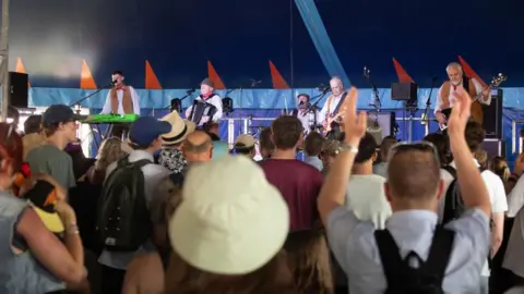 Gorman Photography The photo is taken from amongst the crowd, looking towards the Wurzels performing on stage. One man, with his back to the camera, has his hands in the air as if clapping along with the music.