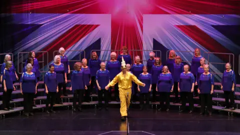 One Acchord A group of around twenty women in blue tops on a stage with a Union Jack backdrop and a man in yellow clothing and a candle-hat in front