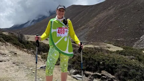 A woman who is dressed as a lime stands in front of a large mountain. She has leggings with pictures of the lime fruit on them plus a bright yellow long-sleeved shift and a top draped across her in green and white slices.
