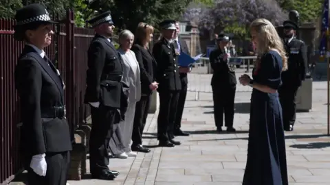 Lissie in a blue dress looking down at the memorial to her late husband, with members of the police stood to attention around her.
