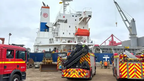 Three fire engines facing a large white ship which is pulled in at a dock. There is a large crane on the right of the ship.