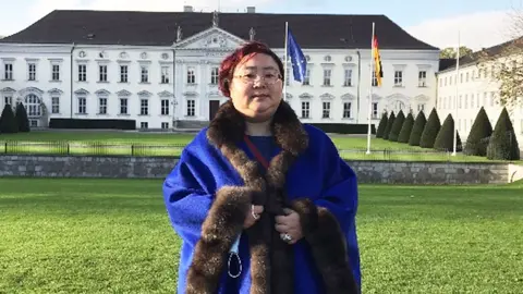 Metropolitan Police Qian Zhemin - wearing glasses and her hair dyed red - standing in front of a building in Berlin. She is wearing a fur-lined bright blue coat. 