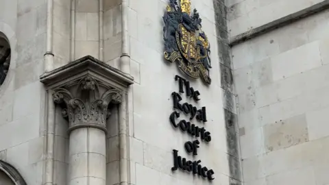 Close up of a sign on a white wall with an emblem featuring a shield, lion and unicorn in black and gold with large black letters beneath proclaiming The Royal Courts of Justice