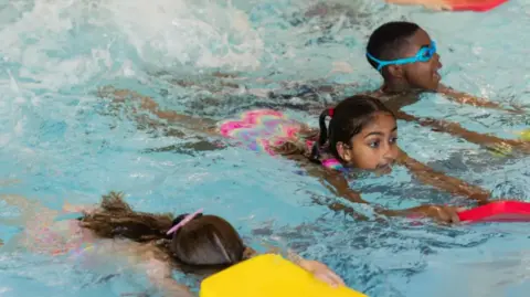AFP via Getty Images Three children swimming with floats in a swimming pool and a swimming lesson.