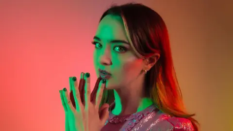 RAPHAEL NEAL Fern Brady posing for a promotional picture, lit in red and green artistic light. She has long dark hair and green eyes. She is wearing a pink sparkly dress and is holding her hands in a prayer gesture under her chin. She is looking sideways at the camera.