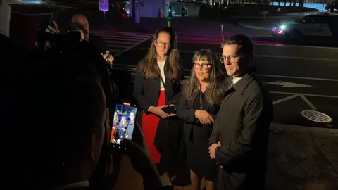 A man and two women stand on a dark street speaking to reporters. The image is dark other than the light from camera shining on the group.