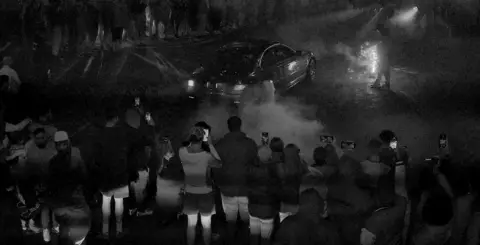 A black and white image of a car meet. There is a group of people all gatherered around a car that has smoke coming from the wheels. 