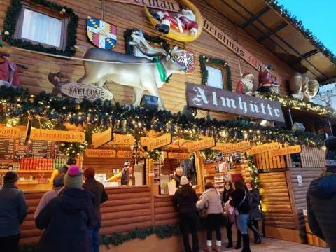 A stall selling alcohol at a Christmas market. The wooden stall is adorned with fake Christmas tree sprigs, golden fairylights, decorations including a giant reindeer and Santa ornaments, signs saying "Almhutte", "Weissbier", "Schneemannpunsch". A group of people in warm winter coats and woolly hats stand in front of the stall, being served by staff