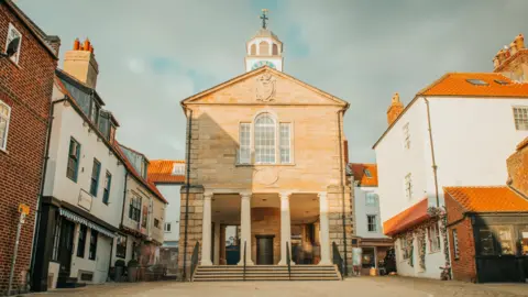 A historic stone building with a columned portico and a small clock tower stands in a sunlit town square, surrounded by traditional red‑roofed buildings and cobblestone paving.