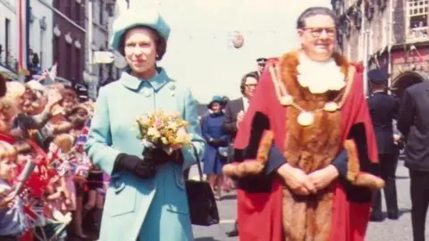 Staffordshire Film Archive A faded colour picture of Queen Elizabeth in a turquoise dress and hat, carrying a bouquet of flowers, walking with dignitaries as she is greeted by crowds.