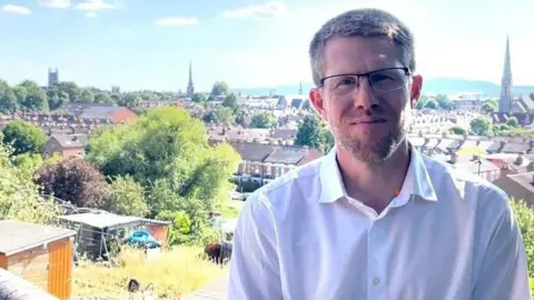 A man wearing a white shirt is standing at the top of a slope - behind him is a view of Worcester city centre, showing allotments, houses, and the top of Worcester Cathedral. 