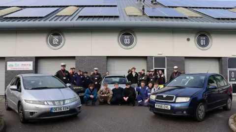 Police and members of the college stand in front of the three donated vehicles. The blue Skoda Fabia is parked on the right and the silver Honda Civic is parked on the left. The silver Citroen C3 is parked in the middle and is surrounded by police and pupils who are looking at the camera. They are stood in front of garage doors. 