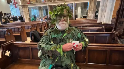 BBC/ Julia Gregory An older man with a white heard is painted in emerald green and wears a multi-shade green costume drinks a cup of tea in a church pew
