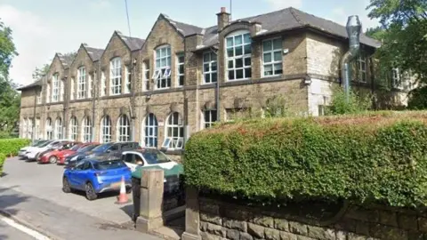 Google Castle Hill Primary School, Todmorden, a building from 1912 with two storeys, a hedge in front and several parked cars. 