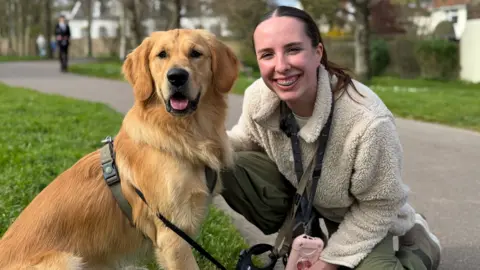 A woman in her 20s with long dark hair kneels on a path in a park beside a golden retriever wearing a harness and lead. There are trees, grass and houses visible in the background.