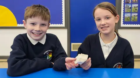 Paul Moseley/BBC Harry and Rosey are smiling at the camera and holding a grey/white piece of RAAC. They're both wearing white polo shirts. Harry has a navy-blue Thomas Bullock Primary jumper over the top his shirt, whilst Rosey is wearing a cardigan in the same colour. 