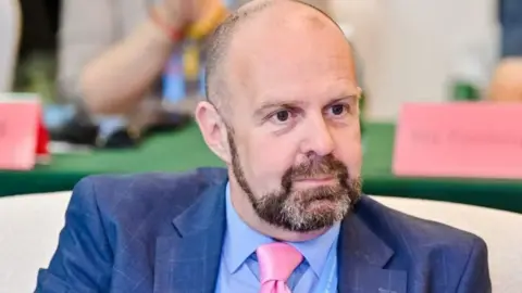 Liverpool City Council regeneration cabinet member Nick Small. He is bald, has a short beard, and is wearing a blue suit, blue shirt and pink tie. 