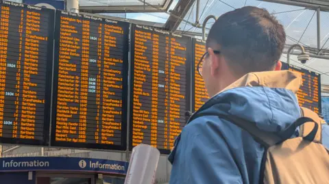 Network Rail A man with short black hair and wearing glasses, a blue jacket and a beige bag is stood in Manchester Piccadilly station looking at the departure board.