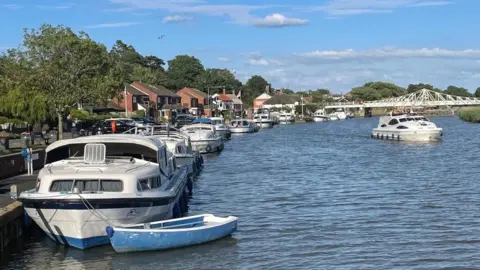 Peter Ward A row of boats moored up at Reedham Quay, with a bridge visible in the background, on a sunny day