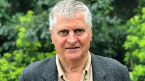 A close up of Rob Prow looking into the camera. He has short grey hair, is wearing a grey jacket and light shirt. He is standing in front of some greenery.