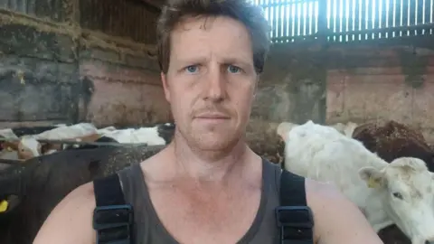 A man with brown hair in a cow shed surrounded by cows