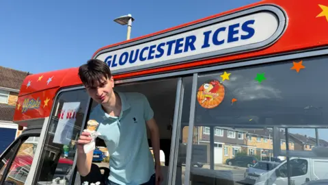 BBC Newsbeat Emery, a young man in a blue polo neck shirt, smiles while holding an ice cream out of the window of an ice cream van, beneath a sign reading: 'Gloucester Ices'