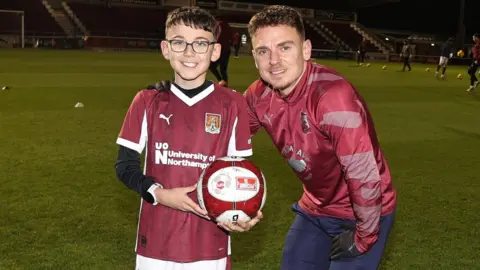 Ezra stands on the pitch next to Sam, who has his arm around Ezra's shoulder, and they both smile at the camera. Ezra holds the football which is in team colours of Maroon and white. He wears the team T-shirt with a University of Northampton logo on, over a black long-sleeved top. The pitch is lit up and there are some players behind them warming up.