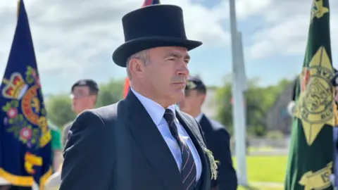 Chief Minister Alfred Cannan in morning dress, including top hat, making his way down the ceremonial walkway, There are standard bearers behind him.