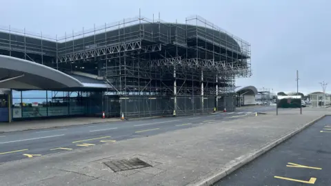 The terminal building covered in scaffolding during construction at Guernsey Airport, with an empty road and bus stop in the foreground under a grey sky.
