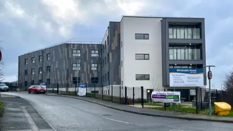 Mark Norman/BBC A large modern grey three storey building with signage outside.